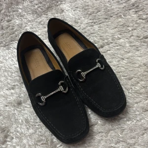 Svarta mockaloafers från Geox - Snygga svarta loafers i mocka från Geox med dekorativ metallspänne på ovansidan. Perfekta för en stilren look. Bekväma och eleganta för olika tillfällen.