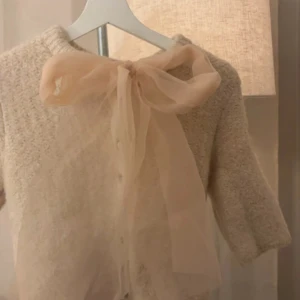 Beige kofta med rosett - Charmig beige kofta med trekvartsärm och en söt rosett framtill. Perfekt för en elegant och feminin look. Koftan har en mjuk och bekväm textur.