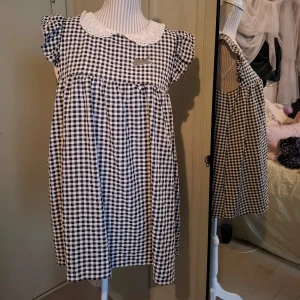 Svartvit rutig klänning med spetskrage - Charmig svartvit rutig klänning med en söt spetskrage och korta ärmar. Klänningen har knappar på baksidan och en lös passform som ger en avslappnad look. Perfekt för en vintageinspirerad stil. Obs det finns ett litet hål i sömmen, går definitivt att fixa