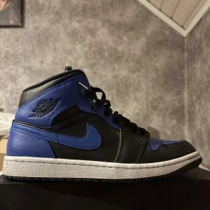 Nike Air Jordan 1 Mid i svart och blå - Säljer ett par Nike Air Jordan 1 Mid i svart och blå. Skorna har en klassisk design med snörning och en hög siluett. Perfekta för dig som älskar sneakers med stil och komfort.
