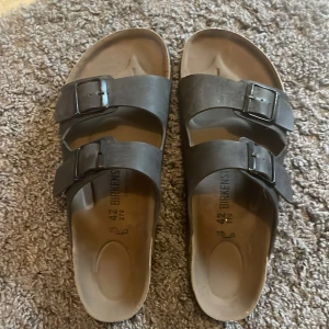 Svarta sandaler från Birkenstock - Säljer ett par klassiska svarta sandaler från Birkenstock med två justerbara remmar och spännen. Perfekta för en avslappnad stil och bekväma att bära hela dagen. Sulan har ett mönster för extra grepp.