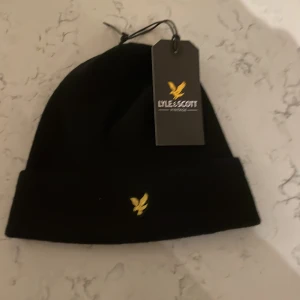 Svart mössa från Lyle & Scott - Snygg svart mössa från Lyle & Scott med deras ikoniska logga i gult framtill. Perfekt för att hålla värmen under kyliga dagar. Enkel och stilren design som passar till det mesta.