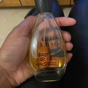 Fabio Verso Exotic Cocktail Eau de Parfum - Säljer en elegant Fabio Verso Exotic Cocktail Eau de Parfum. Flaskan har en stilren, droppformad design med en svart kork. Doften är exotisk och lockande, perfekt för den som vill sticka ut. En perfekt accessoar för att komplettera din stil.
