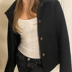 Svart kappa  - Snygg svart kappa från Supercircle med knappar framtill. Den har en klassisk krage och är perfekt för kyligare dagar. Jackan har en stilren design som passar till många olika outfits.