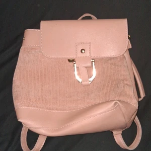 Rosa ryggsäck med manchesterdetaljer - Snygg rosa ryggsäck med en kombination av syntet och manchester. Väskan har ett lock med spänne i metall och justerbara axelremmar. Perfekt för en stilren look.