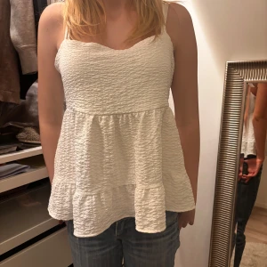 Vit ärmlös topp med volang - Säljer en söt vit ärmlös topp med volangdetaljer. Toppen har en öppen rygg med knytning och en luftig passform. Perfekt för varma dagar!