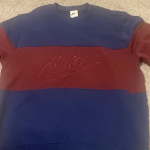 Blå och vinröd tröja från Nike - Säljer en snygg blå och vinröd tröja från Nike med deras ikoniska logga broderad på framsidan. Tröjan har en klassisk passform och är perfekt för en avslappnad stil. Helt ny 