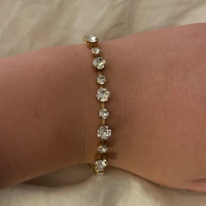 Guldarmband med glittriga stenar - Elegant guldarmband prytt med stora, glittriga stenar. Armbandet har en justerbar kedja och en säker klolås. Perfekt för att ge en touch av glamour till din outfit.