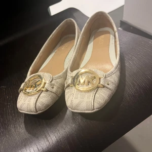 Beige ballerinaskor från Michael Kors - Stilrena beige ballerinaskor från Michael Kors med en elegant gulddetalj framtill. Perfekta för en chic och bekväm look.