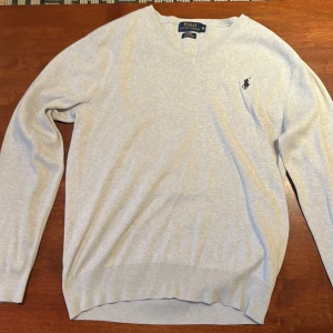 Grå tröja från Polo Ralph Lauren - Säljer en stilren grå tröja från Polo Ralph Lauren i bomull. Tröjan har en v-ringad design och en broderad logotyp på bröstet. Perfekt för en avslappnad men snygg look.