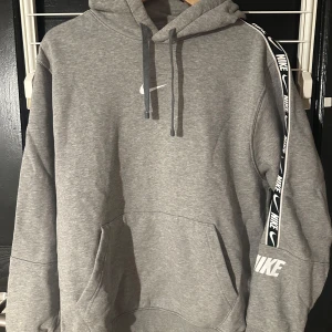 Grå hoodie från Nike - Säljer en grå hoodie från Nike med en klassisk design. Den har en justerbar huva med dragsko och en stor ficka framtill. På ärmen finns ett Nike-band som ger en sportig touch. Perfekt för en avslappnad stil.