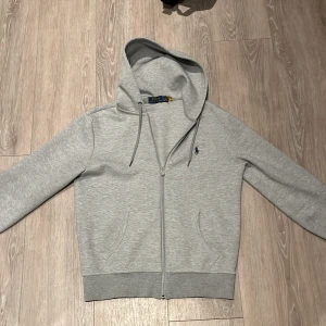 Grå hoodie från Polo Ralph Lauren - Säljer en stilren grå hoodie från Polo Ralph Lauren med dragkedja och huva. Tröjan har långa ärmar och en liten broderad logga på bröstet. Perfekt för en avslappnad look.🤩😍