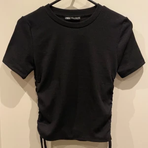 Svart topp från Zara - Snygg svart ribbad topp från Zara med korta ärmar och knytning i sidorna. Perfekt för en stilren look. Toppen har en tight passform och är tillverkad i ett mjukt material och köpt i Spanien men aldrig kommit till användning.  Pris är diskuterat vid snabbt köp ❤️