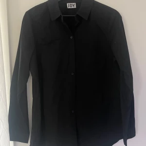 Svart skjorta från JDY - Snygg svart skjorta från JDY med klassisk krage och knappar framtill. Skjortan har långa ärmar och en enkel design som passar till många tillfällen. Storlek 38 men passar en med 36 också. Använd en enstaka gång så i nyskick