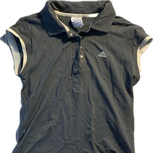 Grå pikétröja från Adidas - Säljer en grå pikétröja från Adidas med korta ärmar och vit kantdetalj. Tröjan har en klassisk krage och knappslå framtill. Perfekt sportig material också!