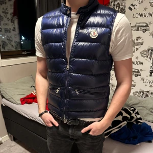 Blå dunväst från Moncler - Säljer en snygg blå dunväst från Moncler med glansig finish. Västen har dragkedja och två praktiska fickor med dragkedjor framtill. Perfekt för att hålla värmen med stil. Lite seg när man drar upp dragkedjan men annars inga defekter! Säljer för den blivit för liten. Pris kan diskuteras! Storlek 1