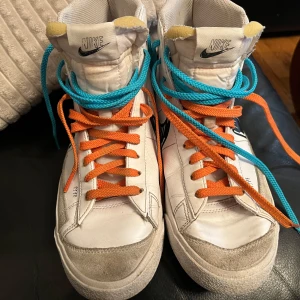 Nike Blazer Mid 77 Black Sketch WhiteReleasing Tomorrow 8 AM!! - Säljer ett par vita Nike sneakers med unika detaljer. Skorna har en hög modell med både orange och blå snörning. De är tillverkade i skinn och har en svart Nike-logga på sidan. Perfekta för en cool och avslappnad stil.