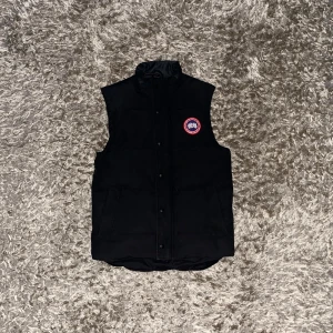 Canada Goose väst - Säljer en svart dunväst från Canada Goose med ett broderat märke på bröstet. Västen har knappar och en hög krage för extra värme. Perfekt för kyliga dagar. Den är storlek XS men sitter bra för någon som har storlek S.