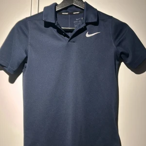 Mörkblå pikétröja från Nike - Säljer en mörkblå pikétröja från Nike med Dri-FIT-teknologi. Tröjan har korta ärmar och en klassisk krage med knappar. Perfekt för sportiga aktiviteter eller en avslappnad stil. Nike-loggan är broderad på bröstet. Fett golf tröja
