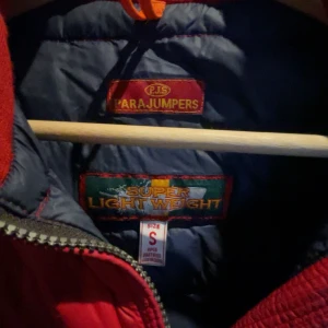 Röd dunjacka från Parajumpers - Snygg röd dunjacka från Parajumpers med quiltad design och huva. Jackan har en dragkedja framtill och den kända patchen på ärmen. Köpt på Vestiarie Collective och är äktighets bevisad av deras experter.