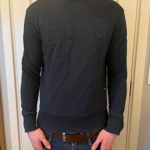Mörkblå tröja från Hugo Boss - Säljer en stilren mörkblå tröja från Hugo Boss med broderad logga på bröstet. Tröjan har långa ärmar och ribbade muddar vid ärmslut och nederkant. Perfekt för en avslappnad och snygg look.