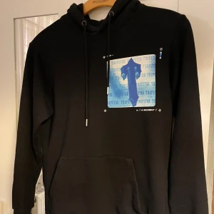 Trapstar Hoodie - Säljer en svart hoodie från Trapstar med unikt blått och vitt tryck på både fram- och baksidan. Hoodien har en klassisk passform med dragsko i huvan och en stor magficka. Perfekt för en avslappnad stil.