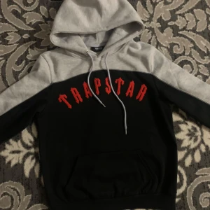 Grå och svart hoodie från Trapstar - Säljer en snygg hoodie från Trapstar i grått och svart med röd text på bröstet. Den har en klassisk huva med dragsko och en stor magficka. Perfekt för en casual look.