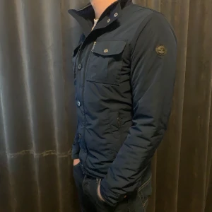  J.Lindeberg field jacket - Snygg mörkblå field jacket från J.Lindeberg med dragkedja och knappar framtill. Jackan har flera praktiska fickor och en stilren design. Perfekt för kyligare dagar.