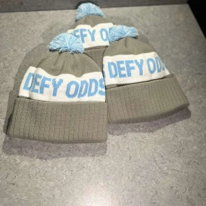 Toppluvan UF signaturmössa ”DEFY ODDS” stötta Barncancerfonden med ditt köp! - Snygg grå mössa med vit och blå text 'DEFY ODDS'. Mössan har en ribbad kant och en ljusblå tofs på toppen. Perfekt för att hålla värmen under kalla dagar.