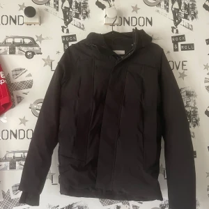 Svart jacka från Jack & Jones - Säljer en stilren svart jacka från Jack & Jones. Jackan har en huva och långa ärmar, perfekt för kyligare dagar. Den är designad med dragkedja och knappar för extra skydd mot väder och vind. Aldrig använd