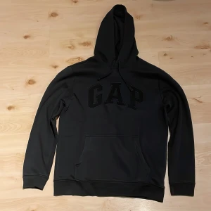 Svart hoodie från GAP - Säljer en stilren svart hoodie från GAP i storlek S. Tröjan har en klassisk design med stor logga på framsidan och en praktisk magficka. Tillverkad i en mjuk bomullsblandning för extra komfort. Perfekt för en avslappnad look.