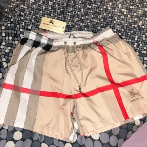 Beige rutig badbyxor från Burberry - Snygga beige badbyxor från Burberry med det klassiska rutmönstret i rött, svart och vitt. De har en elastisk midja med snörning för bästa passform. Perfekta för en stilren look. Endast testade, säljer pågrund att dem sitter lite mer som en Medium🙌