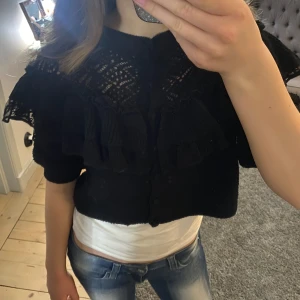 Svart kortärmad kofta med volang - Säljer en svart kofta med korta ärmar och volangdetaljer. Koftan är croppad och har knappar framtill. 