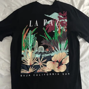 Svart t-shirt med tryck 'La Paz' från Jack & Jones  - Snygg svart t-shirt med färgglatt tryck av växter och texten 'La Paz' på framsidan. Perfekt för en avslappnad stil med en touch av naturinspiration.