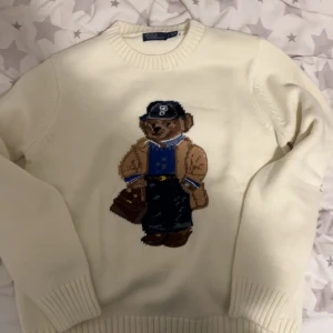Vit tröja med björnmotiv från Polo Ralph Lauren - Säljer en vit tröja från Polo Ralph Lauren med ett coolt björnmotiv på framsidan. Tröjan har långa ärmar och ribbstickade muddar vid ärmslut och nederkant. Perfekt för en avslappnad stil!