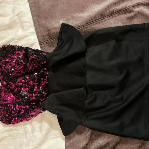 Svart klänning med rosa paljetter stl M - Snygg svart klänning med en överdel täckt av glittriga rosa paljetter. Klänningen är axelbandslös och har en volangdetalj i midjan som ger en extra touch. Perfekt för en kväll ute eller en speciell tillställning.