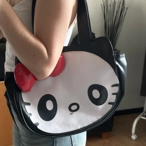  Hello Kitty handväska - Säljer en cool Hello Kitty handväska i svart och vitt med en stor röd rosett. Väskan har ett rymligt rött foder och en praktisk dragkedja. Perfekt för dig som älskar unika accessoarer!