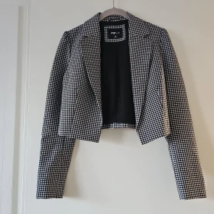 Svartvit rutig kavaj från FB Sister - Snygg svartvit rutig kavaj från FB Sister i storlek M. Kavajen har en croppad design med långa ärmar och är tillverkad i polyester. Perfekt för att ge din outfit en stilren touch.