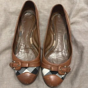 Bruna ballerinaskor med rutigt mönster - Snygga ballerinaskor i brunt skinn med ett klassiskt rutigt mönster i brun, beige, svart. Skorna har en dekorativ spänne framtill som ger en elegant touch. Lite stötta fram vid tån