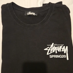 Svart t-shirt från Stüssy - Säljer en svart t-shirt från Stüssy med trycket 'SPRING20' och en färgglad blomdesign på framsidan. T-shirten har korta ärmar och en rund halsringning. Perfekt för en avslappnad stil. Känns mer som L, har bara använt det ett par gånger pga det kalla vädret 