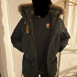 Parajumper herr med päls  - Säljer en svart parkajacka från Parajumpers i storlek M. Jackan har en mysig pälsfodrad huva och praktiska fickor. Perfekt för kalla vinterdagar med sin varma och skyddande design. Den har en dragkedja och knappar för extra skydd mot kyla. Passar både till vardags och mer äventyrliga utflykter.