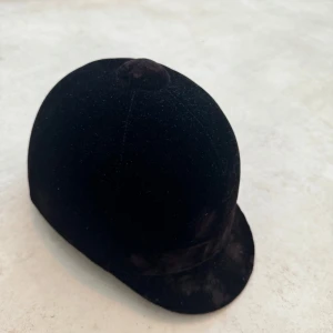 Svart ridhjälm från CRW - Säljer en svart ridhjälm från CRW. Hjälmen har en klassisk design med en mjuk yta och en gul insida. Den är utrustad med justerbara remmar för en säker passform. Perfekt för ridning och i bra skick!