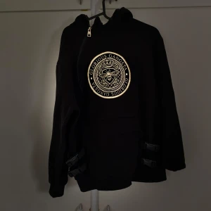 Svart hoodie från Glorious Gangsta - Säljer en svart hoodie från Glorious Gangsta med ett stort, coolt tryck i guld på framsidan. Den har en dragkedja vid halsen och är perfekt för en avslappnad stil. Passar bra till både vardag och chillkvällar. Hör av dig om du är intresserad!