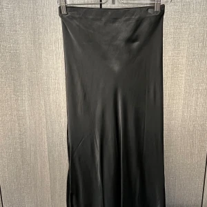 Svart långkjol i satin - Snygg svart långkjol i satin som passar perfekt för både fest och nyår. Kjolen har en elegant och glansig yta som ger en lyxig känsla. Den är i nyskick och har en normal passform. Perfekt för att styla med en glittrig topp eller en enkel blus.