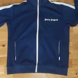 Blå träningsjacka från Palm Angels - Säljer en snygg blå träningsjacka från Palm Angels. Den har vita detaljer och dragkedja framtill. Jackan är i polyester och har en cool, sportig stil med långa ärmar och vita ränder längs sidorna. Perfekt för både träning och vardagsbruk!