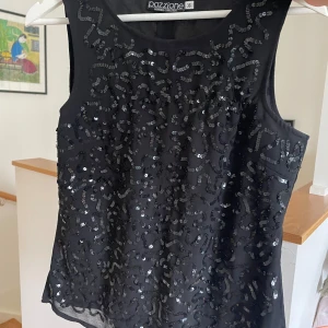 Svart paljettprydd topp från Pazzione - Snygg svart ärmlös topp från Pazzione med glittrande paljetter över hela framsidan. Perfekt för festliga tillfällen! Toppen är i storlek 36 och har en elegant och stilren design. Baksidan är enkel och utan paljetter.
