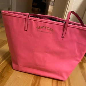 Rosa shoppingväska  - Säljer en superfin rosa shoppingväska med texten 'NEW YORK I THINK I LOVE YOU' på framsidan. Väskan har ett coolt mönster inuti i svart, vitt och rosa. Perfekt för en dag på stan eller som en snygg strandväska. Den är rymlig och har bekväma handtag.