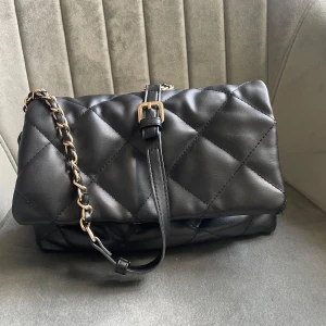 Svart quiltad axelväska - Snygg svart quiltad axelväska i skinn med kedjedetaljer på axelremmen. Väskan har en praktisk storlek med flera fack inuti för enkel organisering. Perfekt för både vardag och festliga tillfällen. Den har en stilren design med en justerbar rem och en säker stängning.