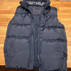 Säljer en snygg svart dunväst från Tommy Hilfiger. Västen har en dragkedja framtill och är perfekt för kyliga dagar. Den är quiltad och har en klassisk stil som passar både höst och vinter. Aldrig använd ny pris 1000, pris går att förhandlas. 