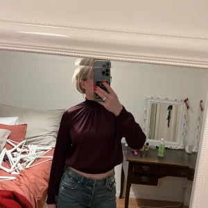 Vinröd blus med hög krage - Säljer en elegant vinröd blus med hög krage och långa ärmar. Blusen har en fin rynkad detalj framtill och en knappöppning i nacken. Perfekt för höst och vinter, både till vardags och fest. Passar bra med jeans eller kjol